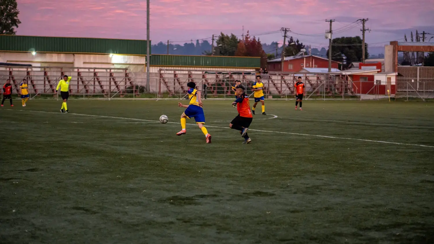 Partido en la cancha al atardecer, con el cielo rosado sobre el pueblo.